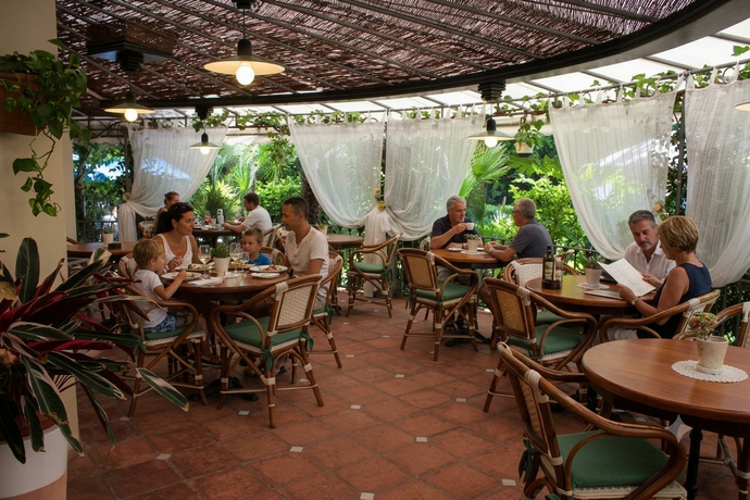 Terrazza all'aperto con tavoli e sedie in legno, tende bianche e una vegetazione rigogliosa.