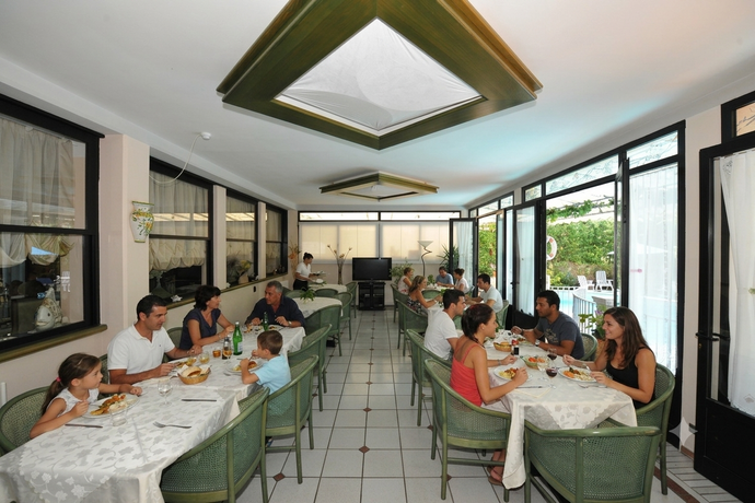 Luminosa sala da pranzo con sedie verdi, tovaglie bianche e grandi finestre che si affacciano su un giardino.