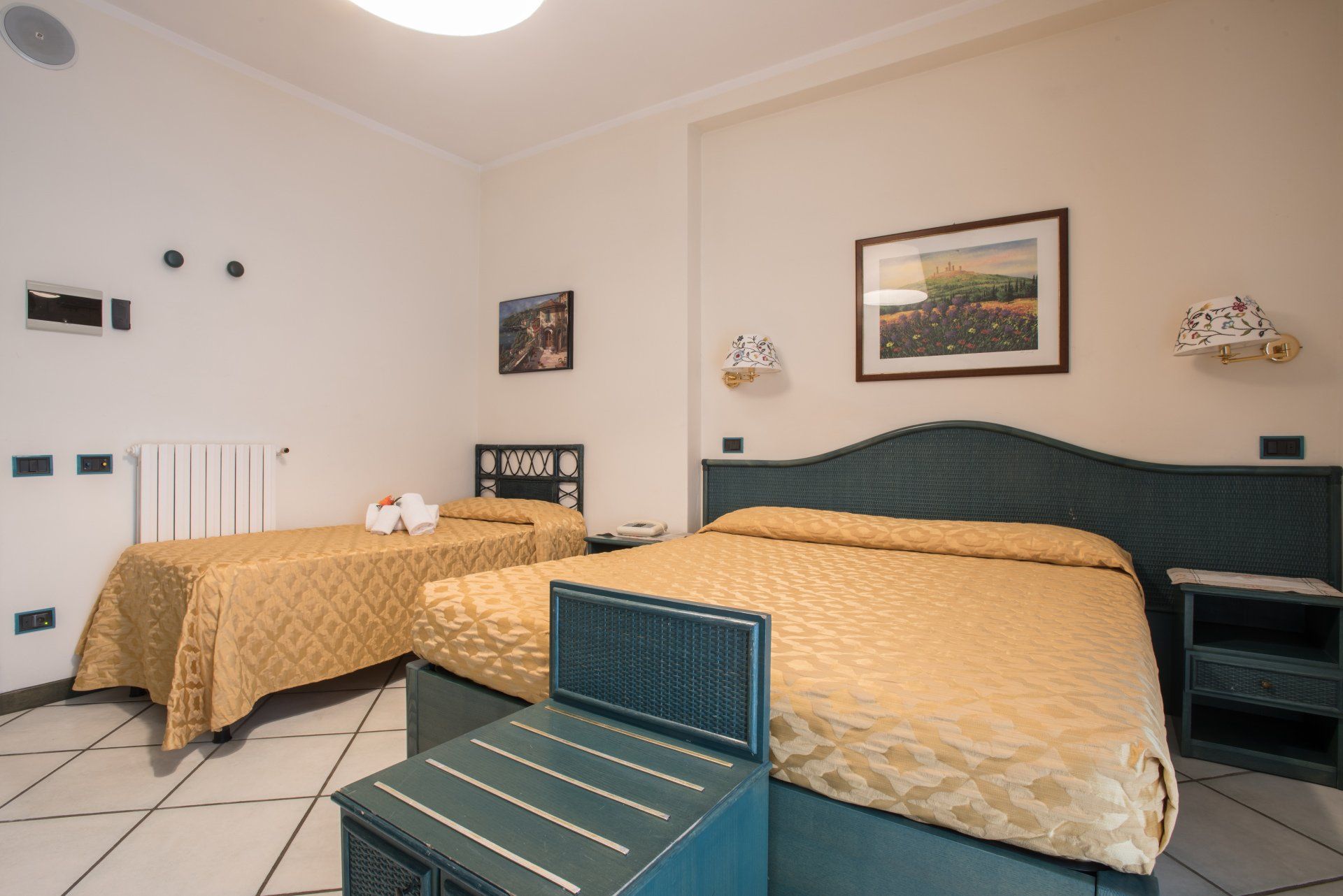 Camera d'albergo con tre letti, biancheria da letto beige e quadri alle pareti in una stanza piastrellata.