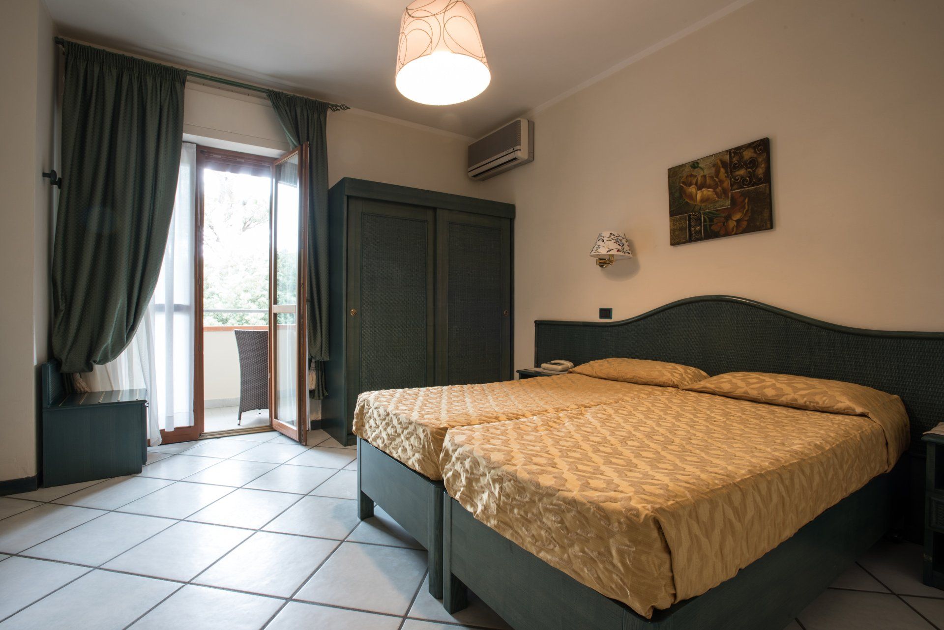Camera da letto con un grande letto, pavimento piastrellato, tende scure e porta-finestra che dà sul balcone e lascia entrare la luce naturale.