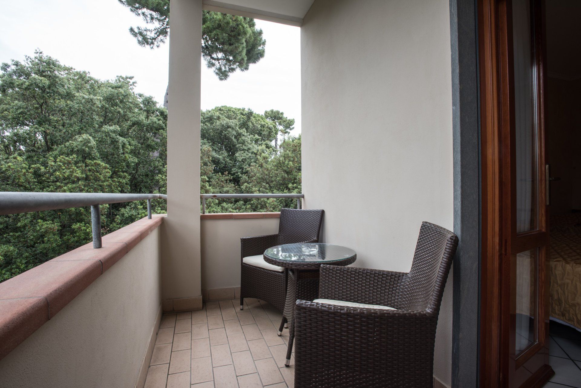 Balcone con sedie in vimini e tavolo in vetro con vista su alberi verdi.