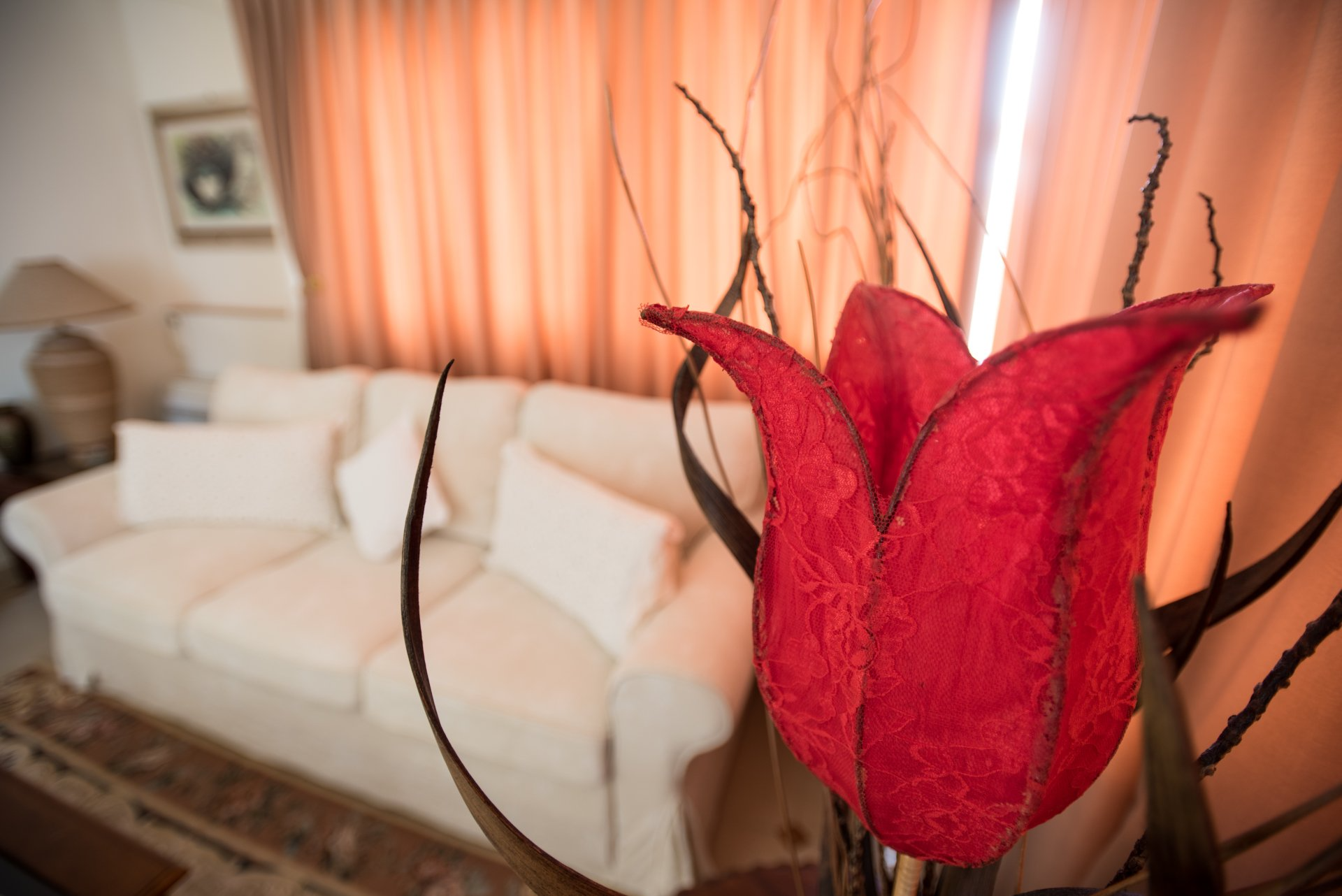 Un fiore decorativo rosso davanti a un divano bianco e tende color pesca in un soggiorno.