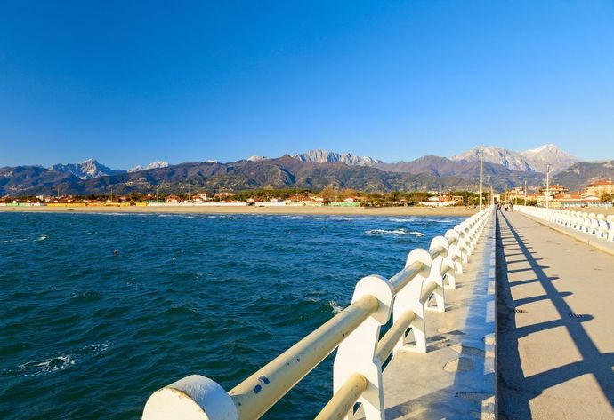 Passeggiata soleggiata sul lungomare con acqua azzurra, montagne sullo sfondo e una lunga ringhiera bianca che proietta ombre