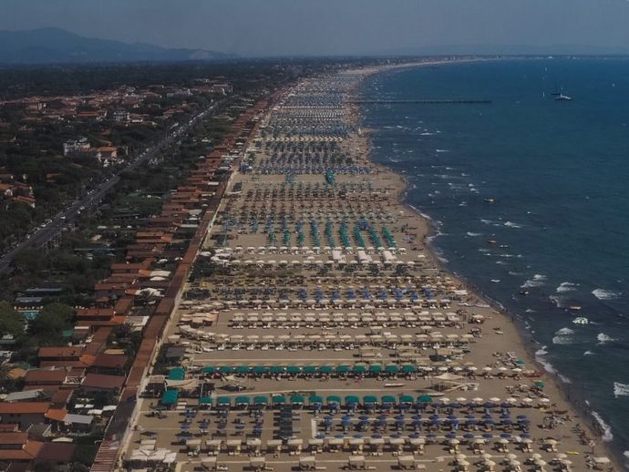 Veduta aerea di una spiaggia affollata, fiancheggiata da ombrelloni, accanto a una cittadina costiera e al mare blu.