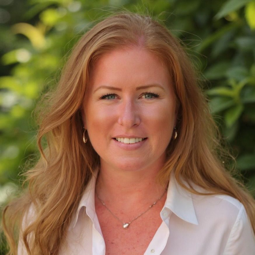 Woman with long red hair smiles, wearing a white shirt and gold jewelry, set against a green leafy background.