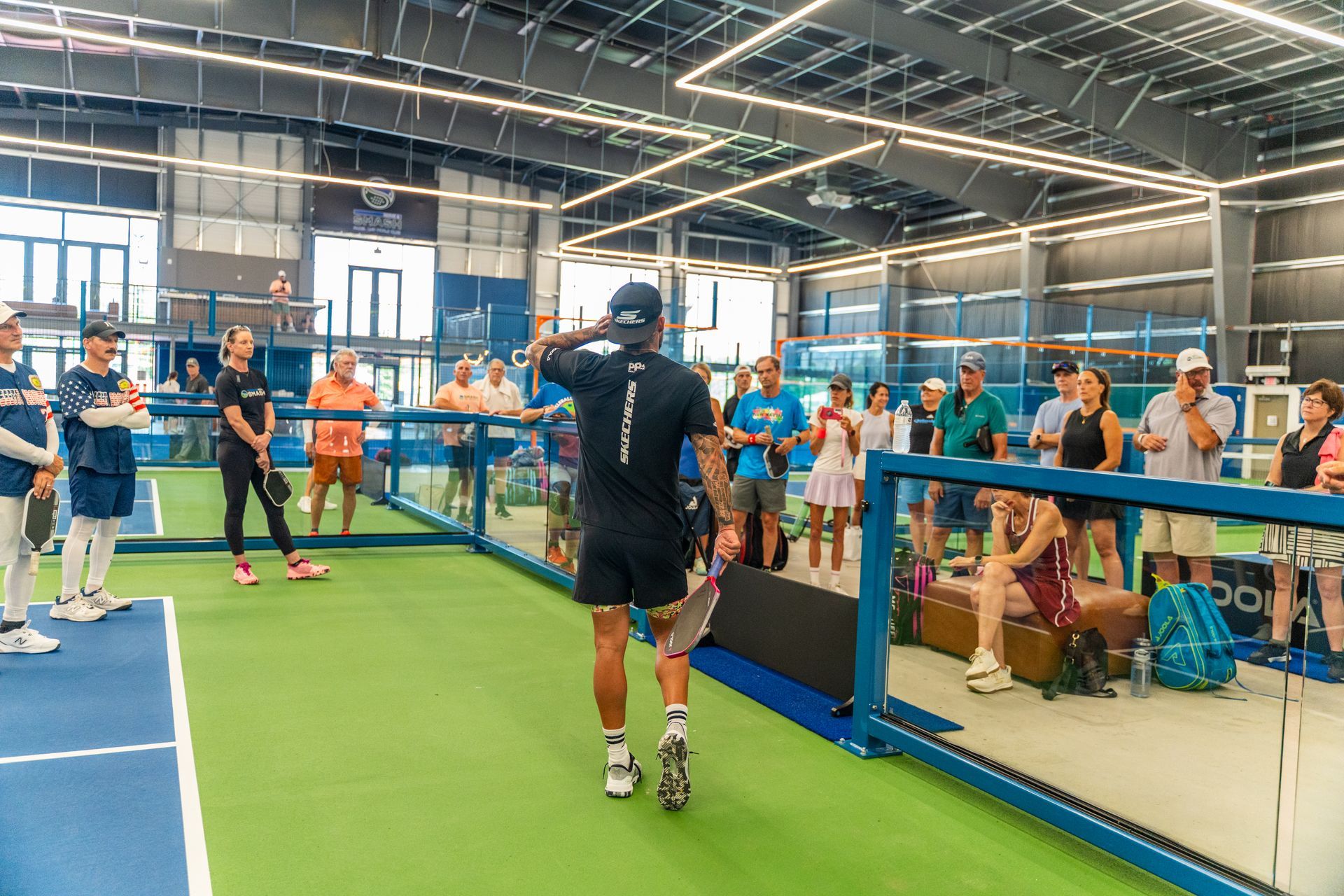 Man in black facing a crowd on a pickleball court, gesturing while people watch.