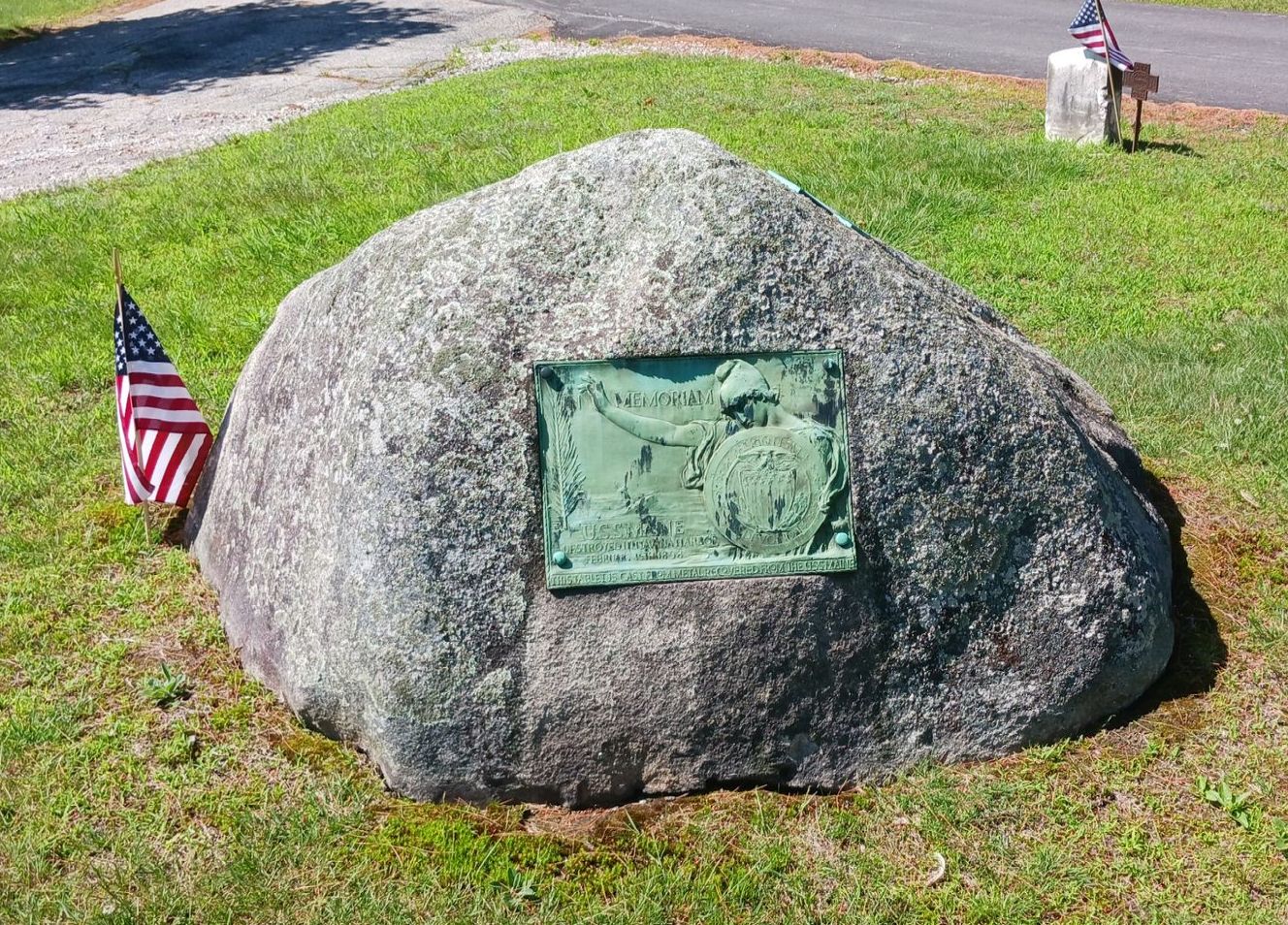 A weathered stone monument with a bronze plaque of figures, set in green grass, with small American flags on each side.