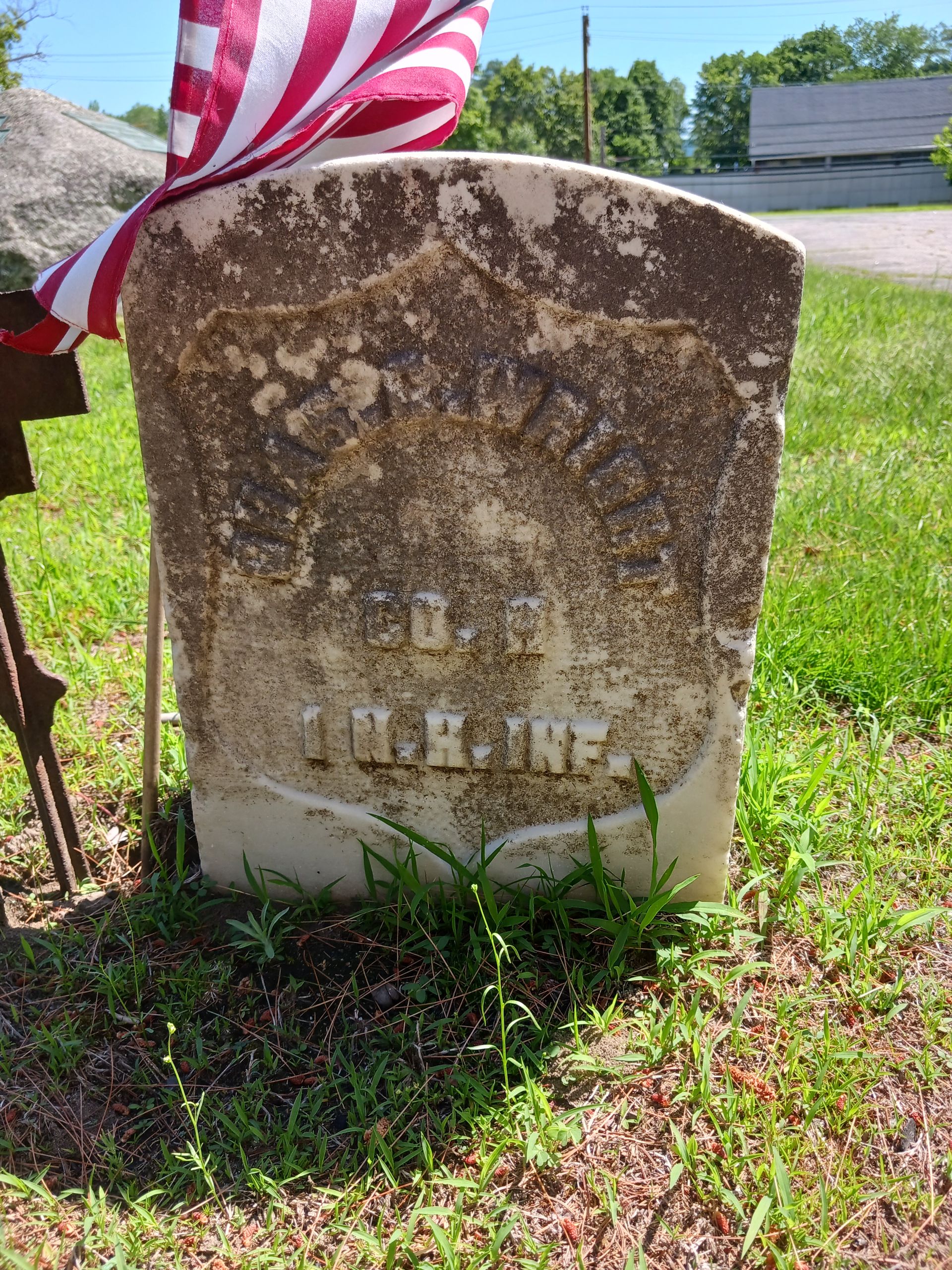 Gravestone with worn inscription, partially obscured by age, in a grassy area; American flag leans on the stone.