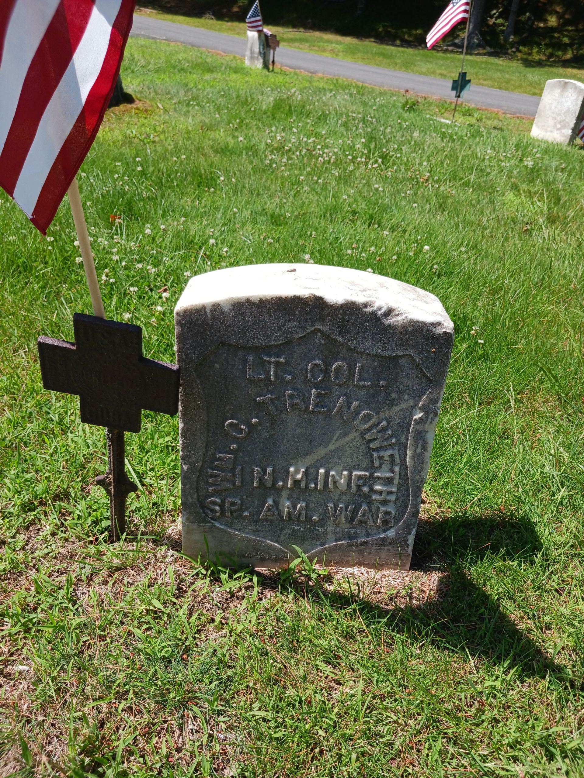 Grave marker in a grassy cemetery with an American flag and a cross.