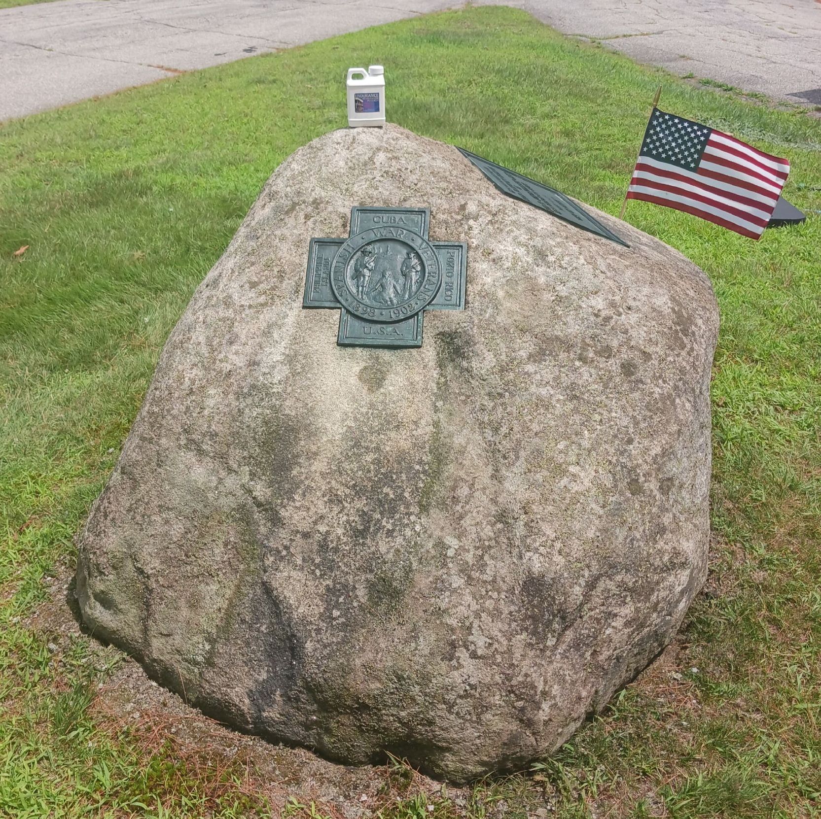 Large stone monument with a bronze plaque and an American flag. Located on grass.