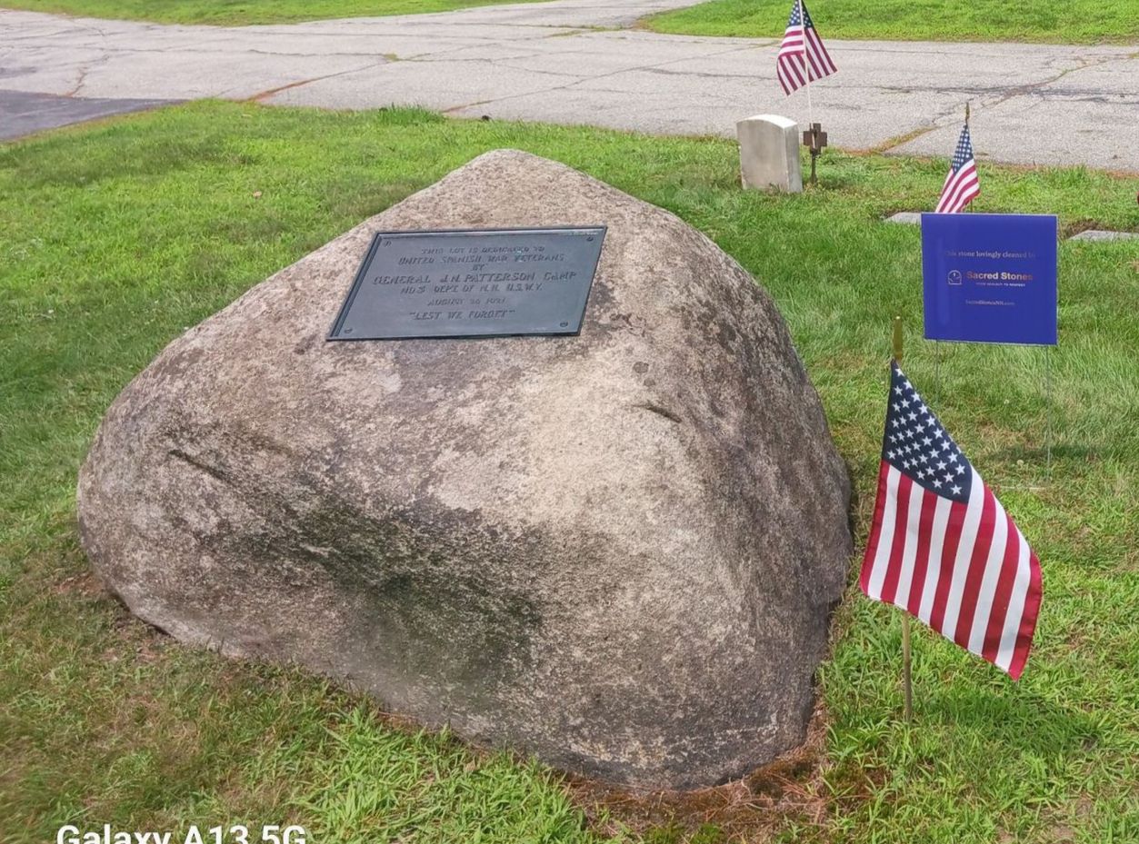A large, gray boulder with a plaque in a grassy cemetery, decorated with American flags.