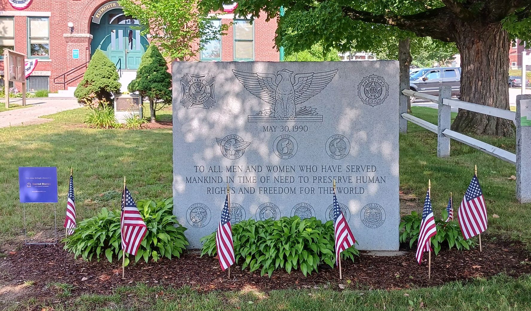 Memorial with American flags in front. The gray stone monument has engraved symbols and text honoring veterans.