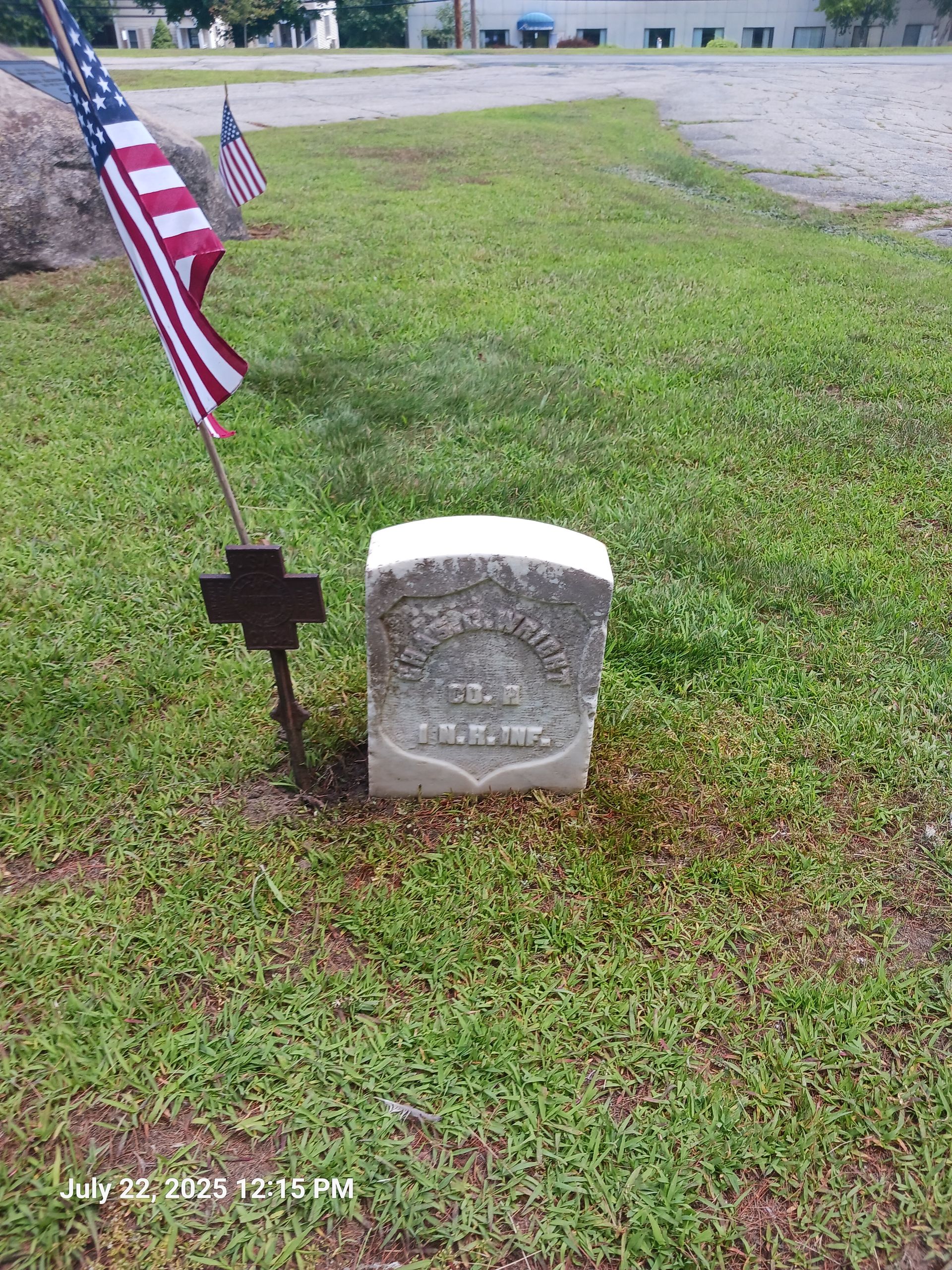 A tombstone in a grassy area, flanked by American flags and a black cross. The headstone is white with engraved text.
