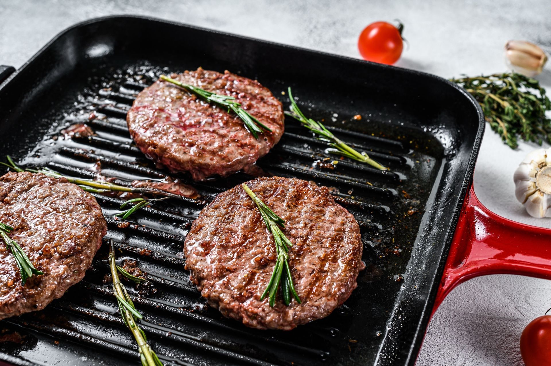 Three grilled hamburger patties with rosemary on a black grill pan.