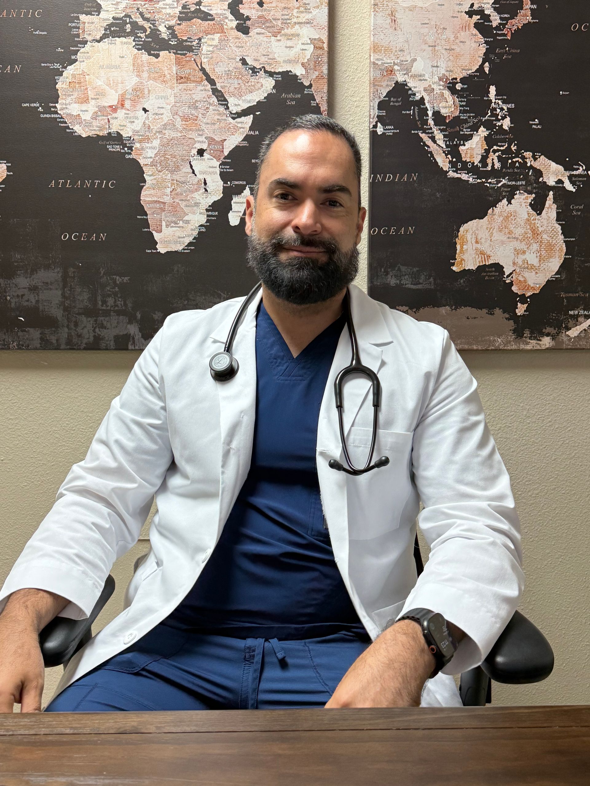 Doctor in white coat and navy scrubs seated before a world map wall, wearing a stethoscope.