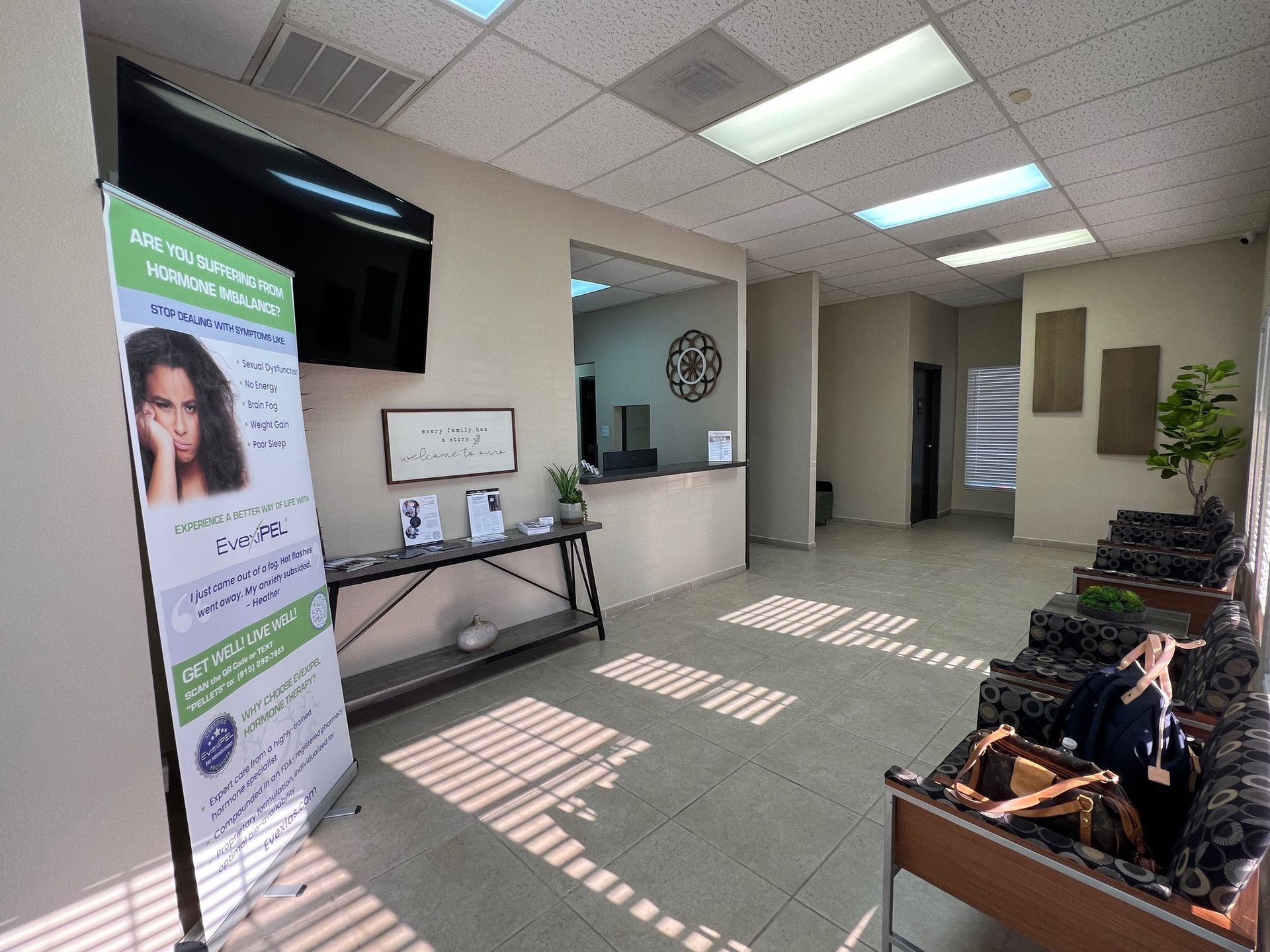 Waiting room with a banner, table, chairs, and sunlit floor tiles in a clinic or office lobby.