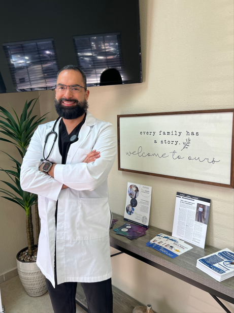 Doctor in white coat standing with arms crossed in a clinic lobby beside a framed quote and brochures