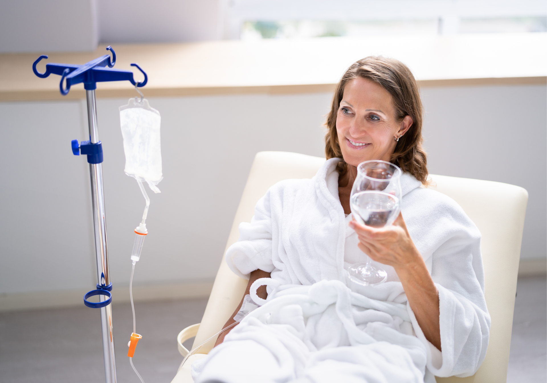 Woman in a white robe sitting in a chair, holding a glass beside an IV stand in a bright room