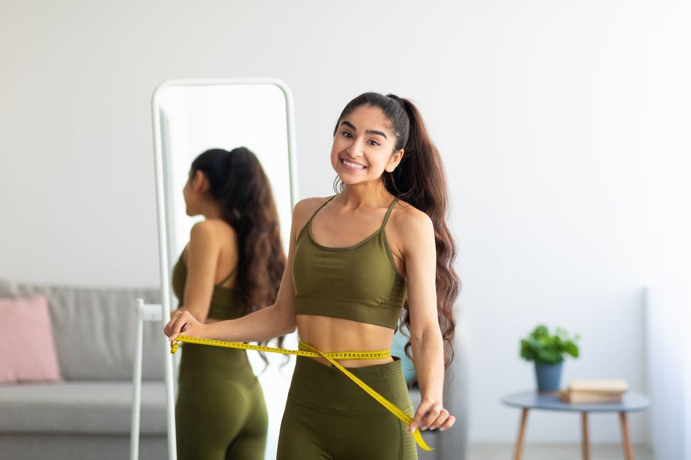 Woman measuring her waist with a tape measure in a bright room, standing beside a full-length mirror.