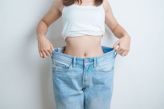 Woman holding oversized blue jeans around her waist against a plain background
