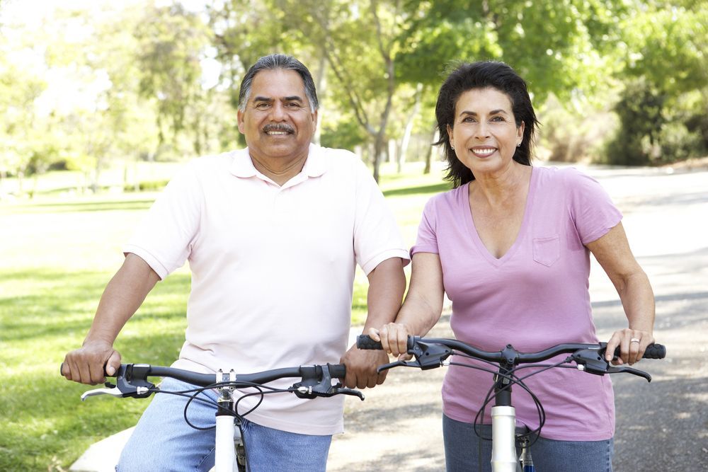Two smiling cyclists holding handlebars in a sunny park, with trees and grass in the background.