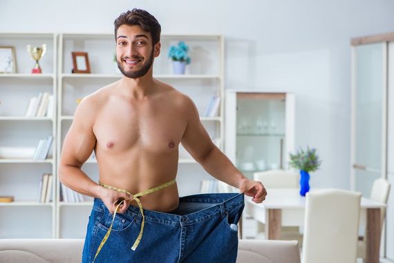 Smiling man holding a measuring tape around his waist, wearing oversized jeans in a bright room