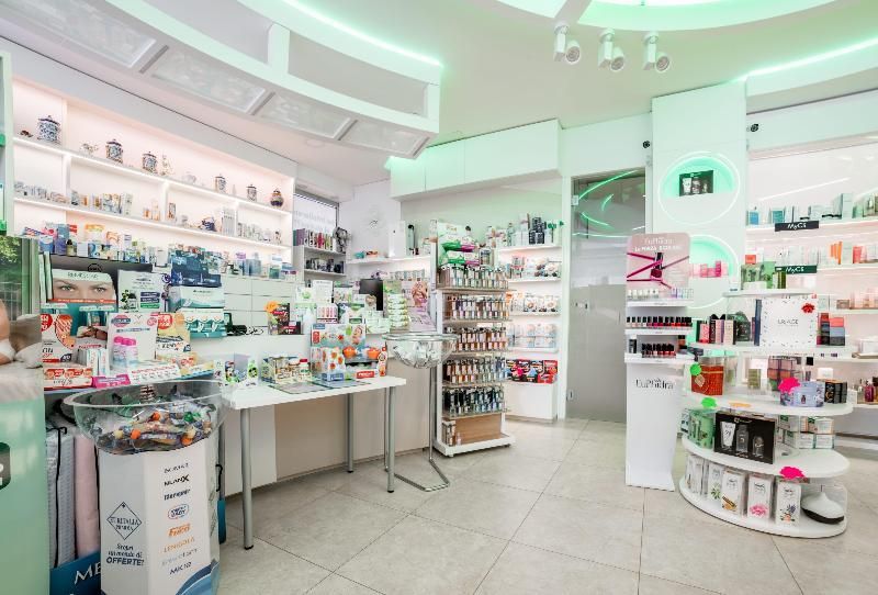 Interno luminoso e bianco di un negozio di prodotti di bellezza e cosmetici con scaffali, tavoli espositivi e prodotti sparsi ovunque.