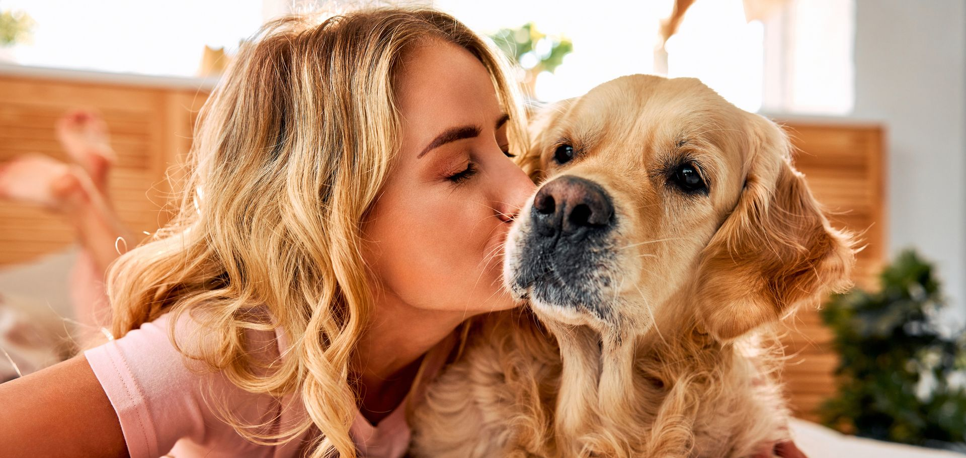 Una donna bacia un golden retriever accanto a una finestra luminosa, entrambi in un ambiente interno accogliente.