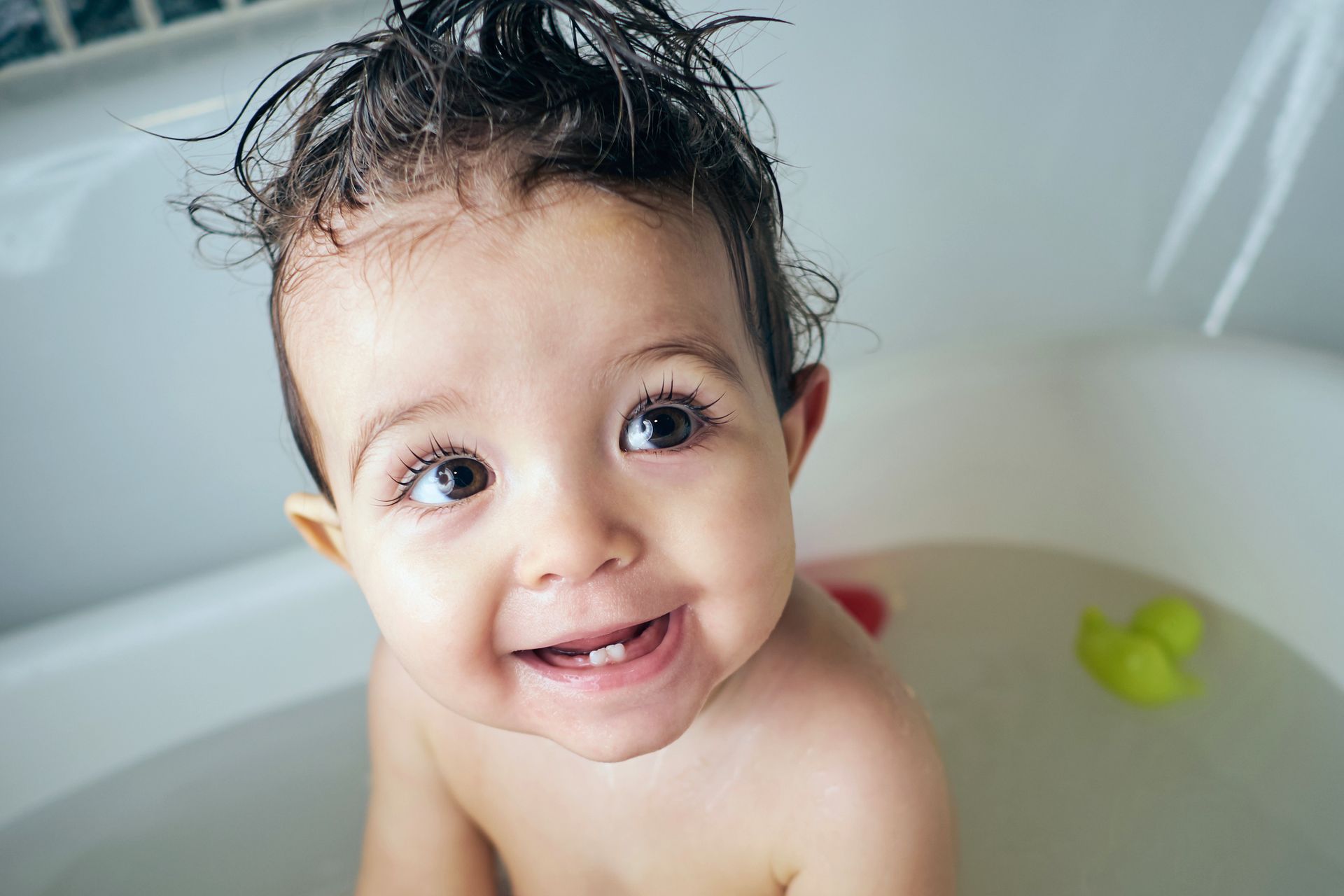 Bambina sorridente nella vasca da bagno con i capelli bagnati e un piccolo giocattolo giallo nelle vicinanze.