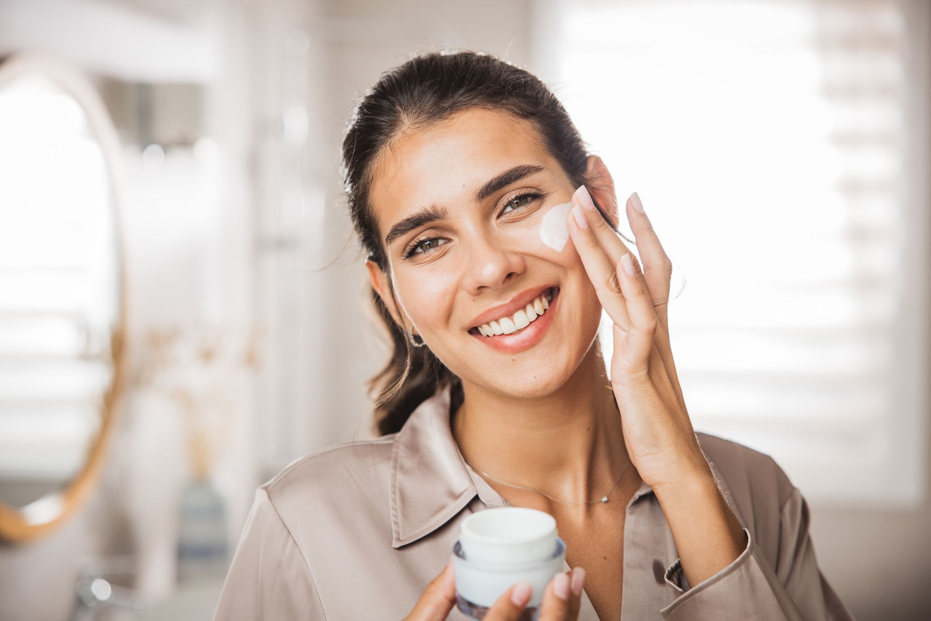 Una donna sorridente si applica una crema per il viso in un bagno luminoso, tenendo in mano un vasetto di prodotti per la cura della pelle.
