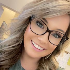 Close-up portrait of a smiling person with blonde, shoulder-length wavy hair wearing dark-rimmed eyeglasses.
