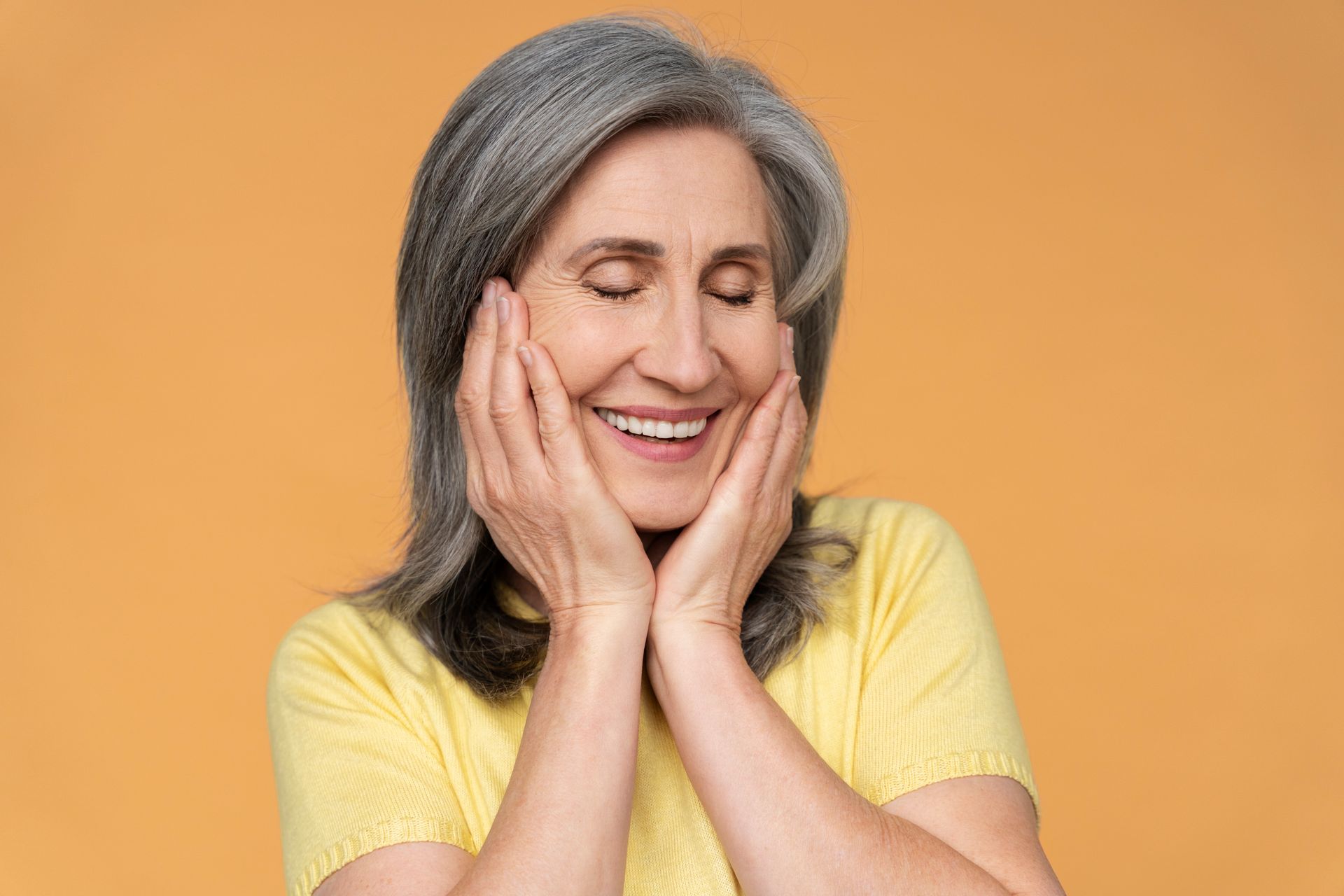 An elderly woman is smiling with her eyes closed and her hands on her face.