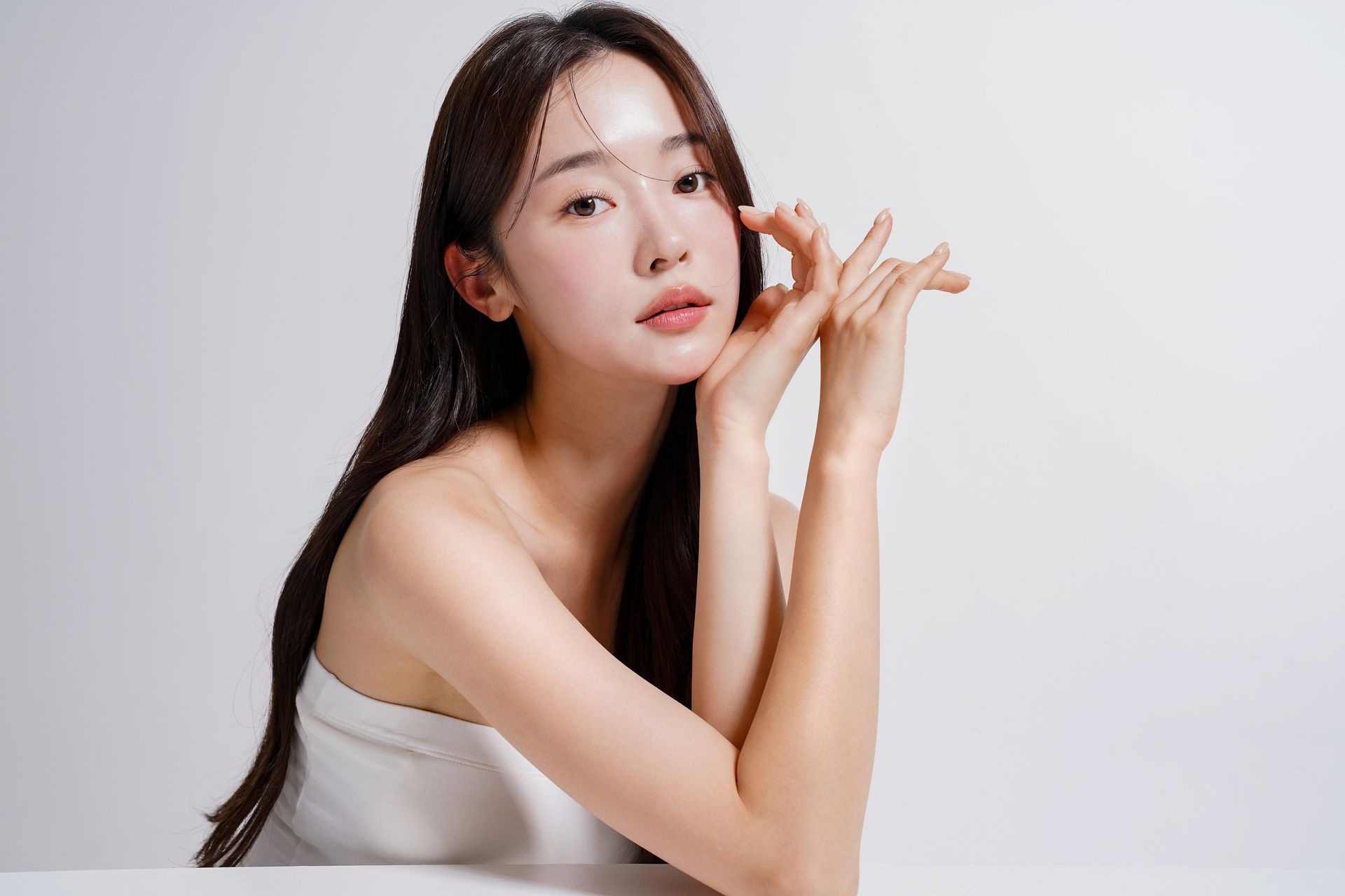 Woman posing in a white top against a light background, resting her hands near her face.