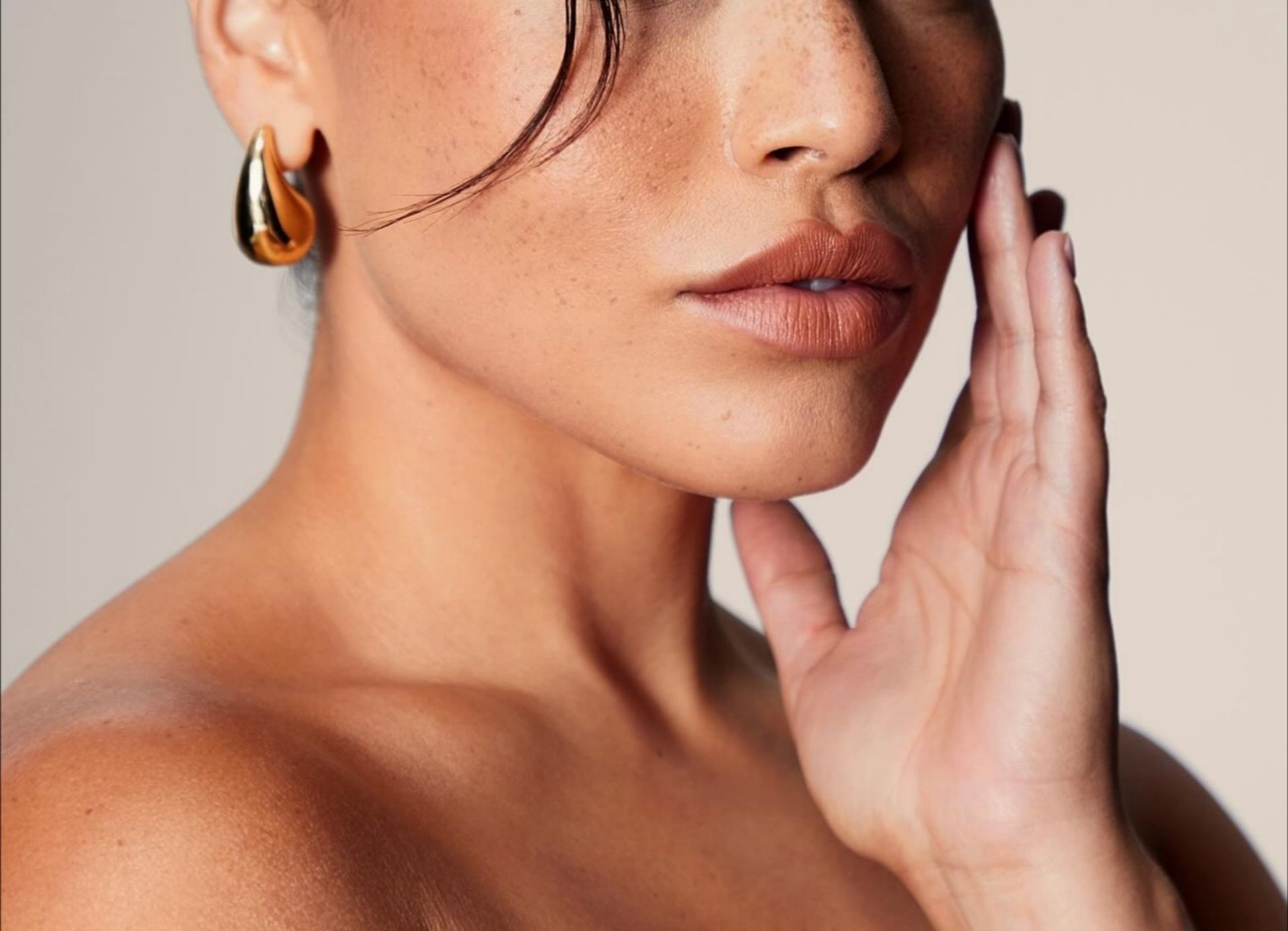 Woman with golden hoop earrings touches her cheek; sun-kissed skin, neutral background.