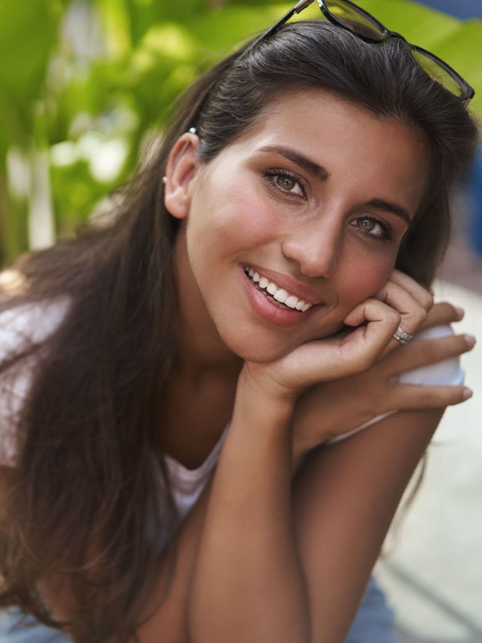 A woman wearing sunglasses is smiling with her hand on her chin.
