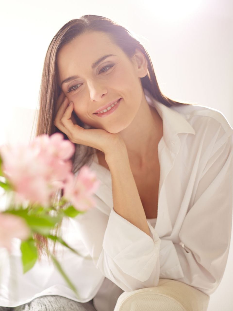 A woman in a white shirt is sitting in front of a vase of pink flowers.