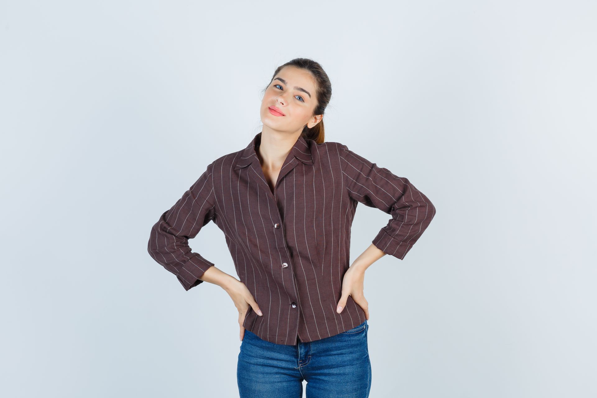 A woman in a brown shirt and blue jeans is standing with her hands on her hips.