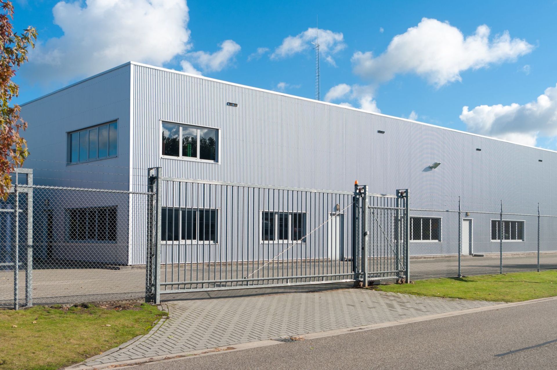 A silver, industrial warehouse building with a chain-link fence and a sliding gate in front under a blue, cloudy sky.