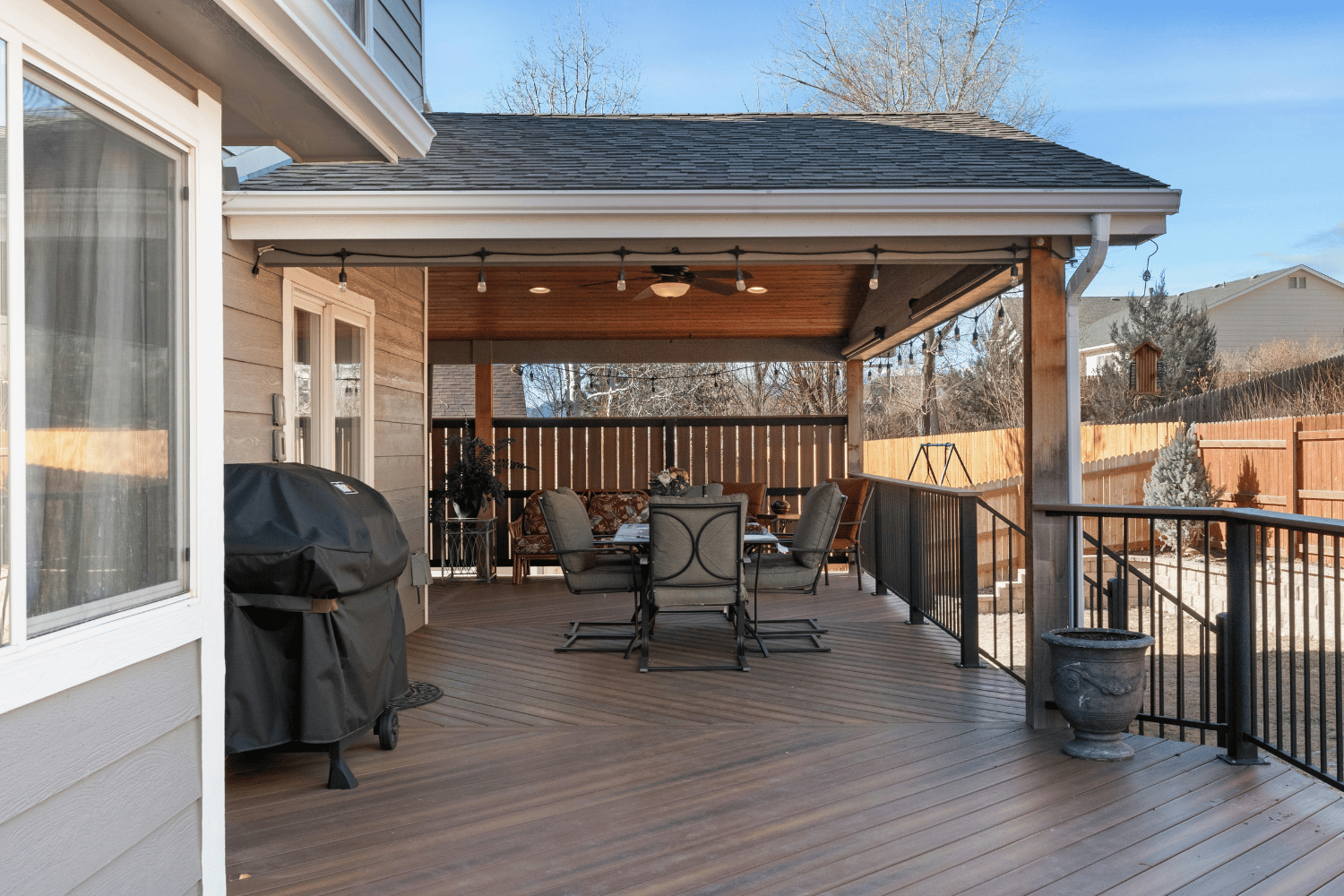 Covered wooden deck with dining table, grill, and railing overlooking a snowy backyard and hills