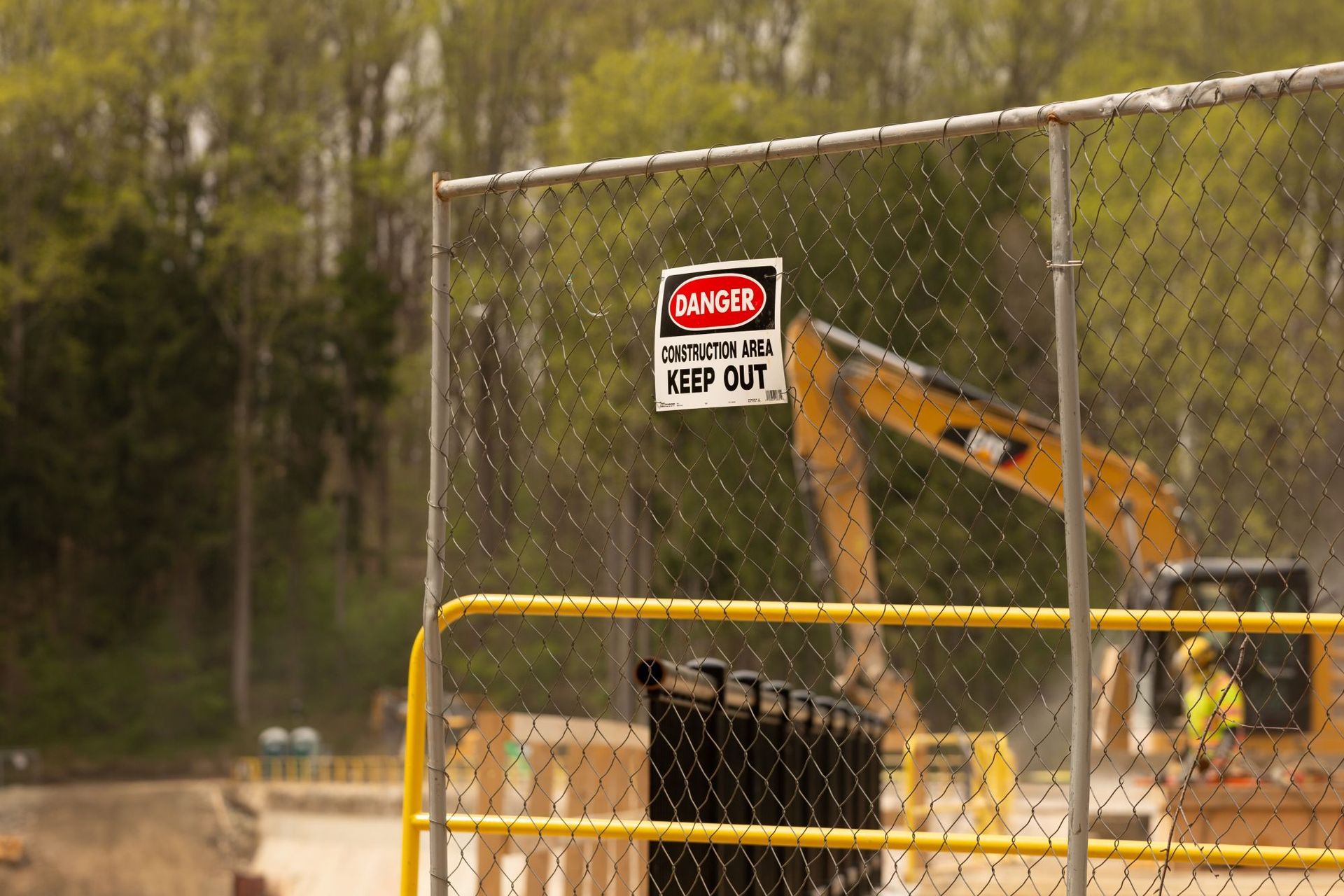 A construction area warning sign on a chain-link fence, with a yellow excavator working in the background.