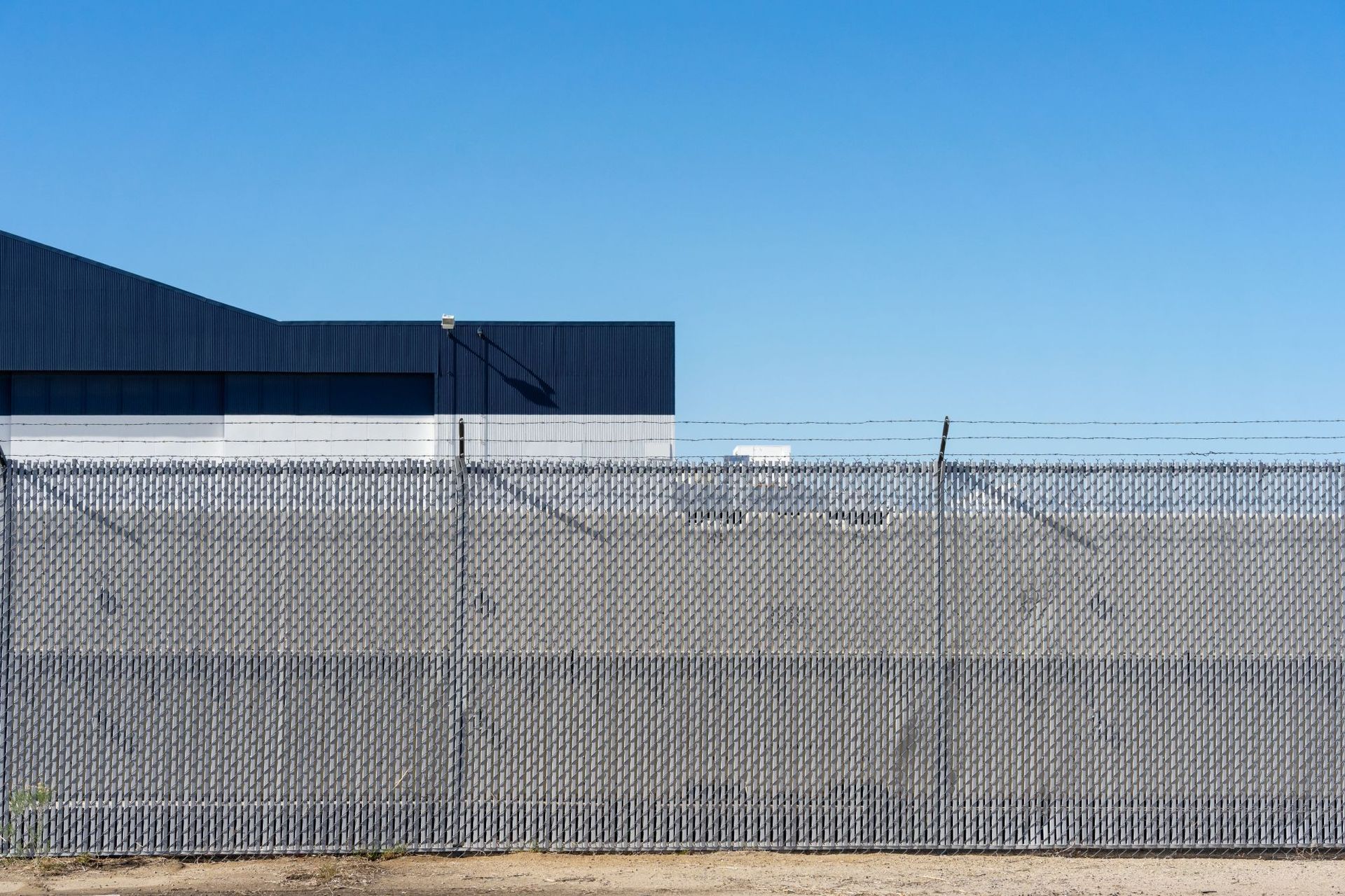 A chain-link fence topped with barbed wire stands in front of a modern industrial building against a clear blue sky.