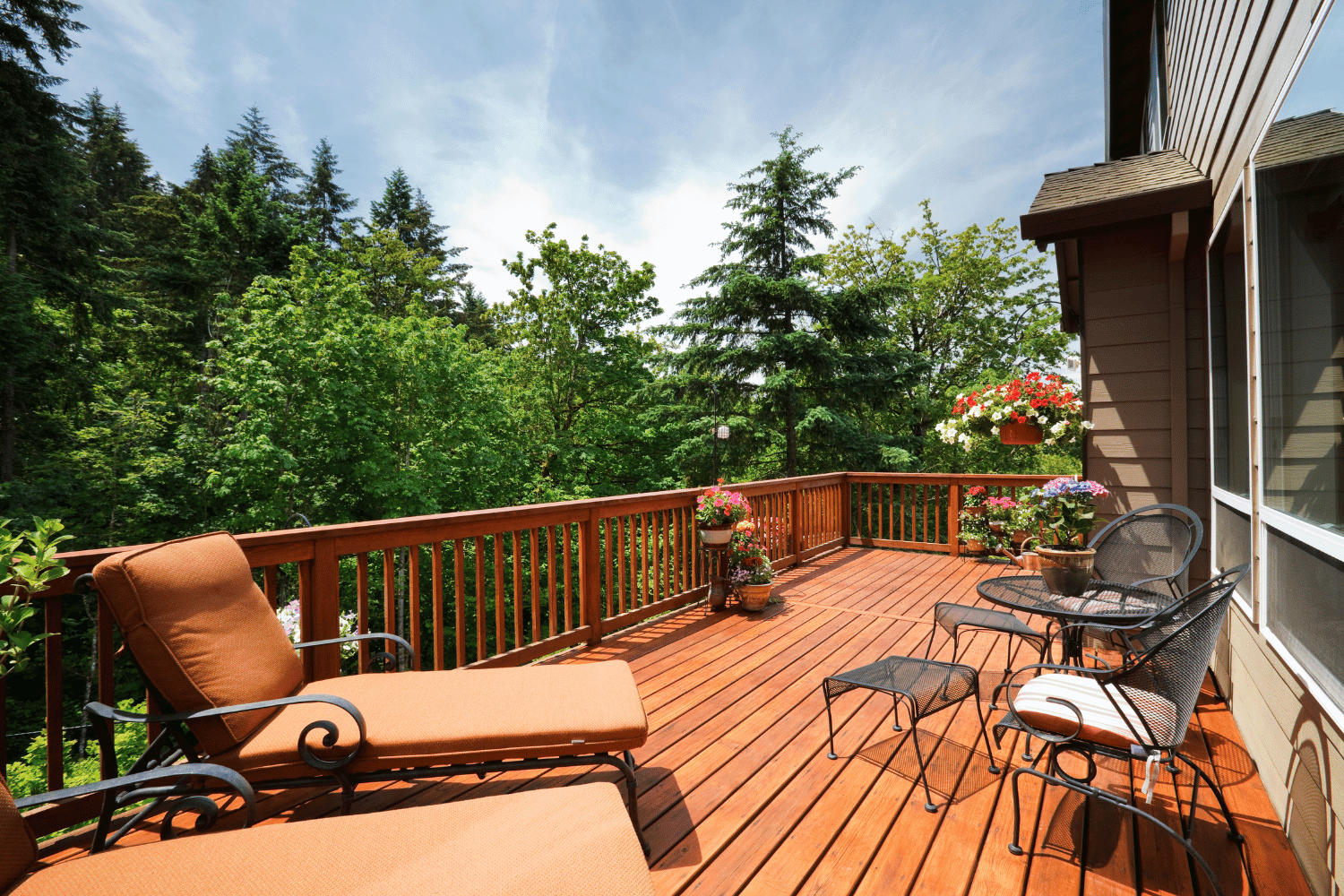 A wood deck overlooking a lush green forest, featuring orange lounge chairs and a small table with chairs and flowers.