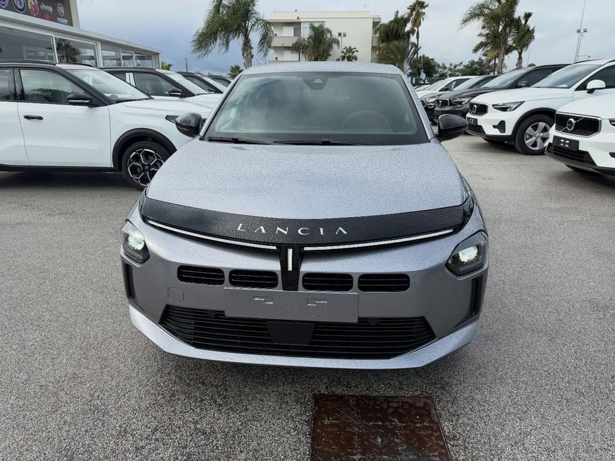 Lancia argentata parcheggiata all'aperto con altre auto. Scritta LANCIA sul cofano. Paraurti grigio e finiture nere.