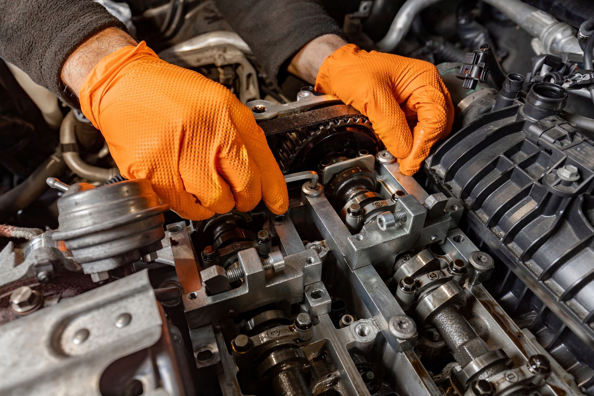 Orange-gloved hands repair an exposed car engine with tools and components visible.