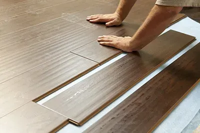 A pair of hands installs dark wood-look laminate floor planks over a light-colored underlayment.