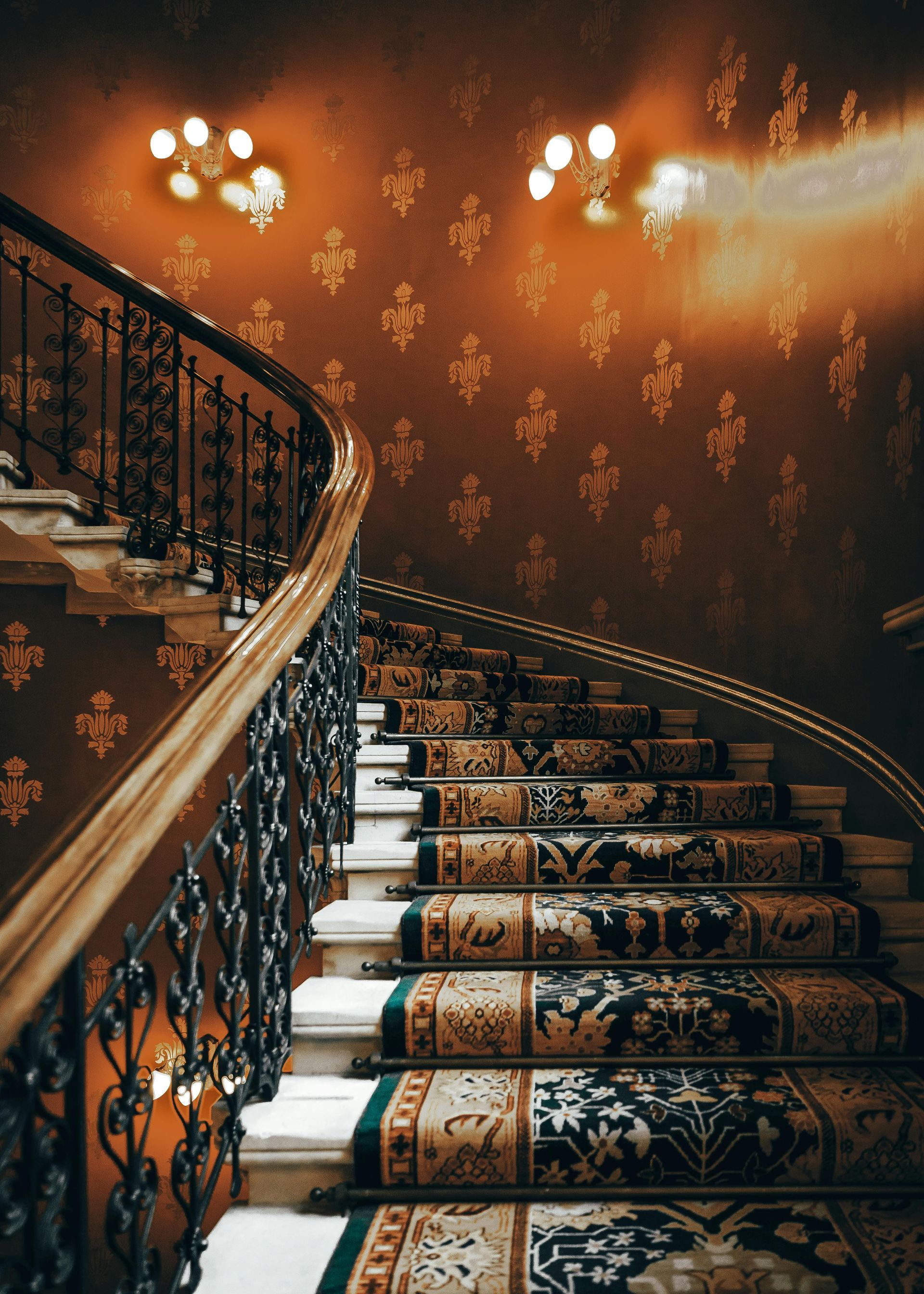 A grand, curved staircase with ornate iron railings, patterned carpet, and light fixtures against a floral-patterned wall.