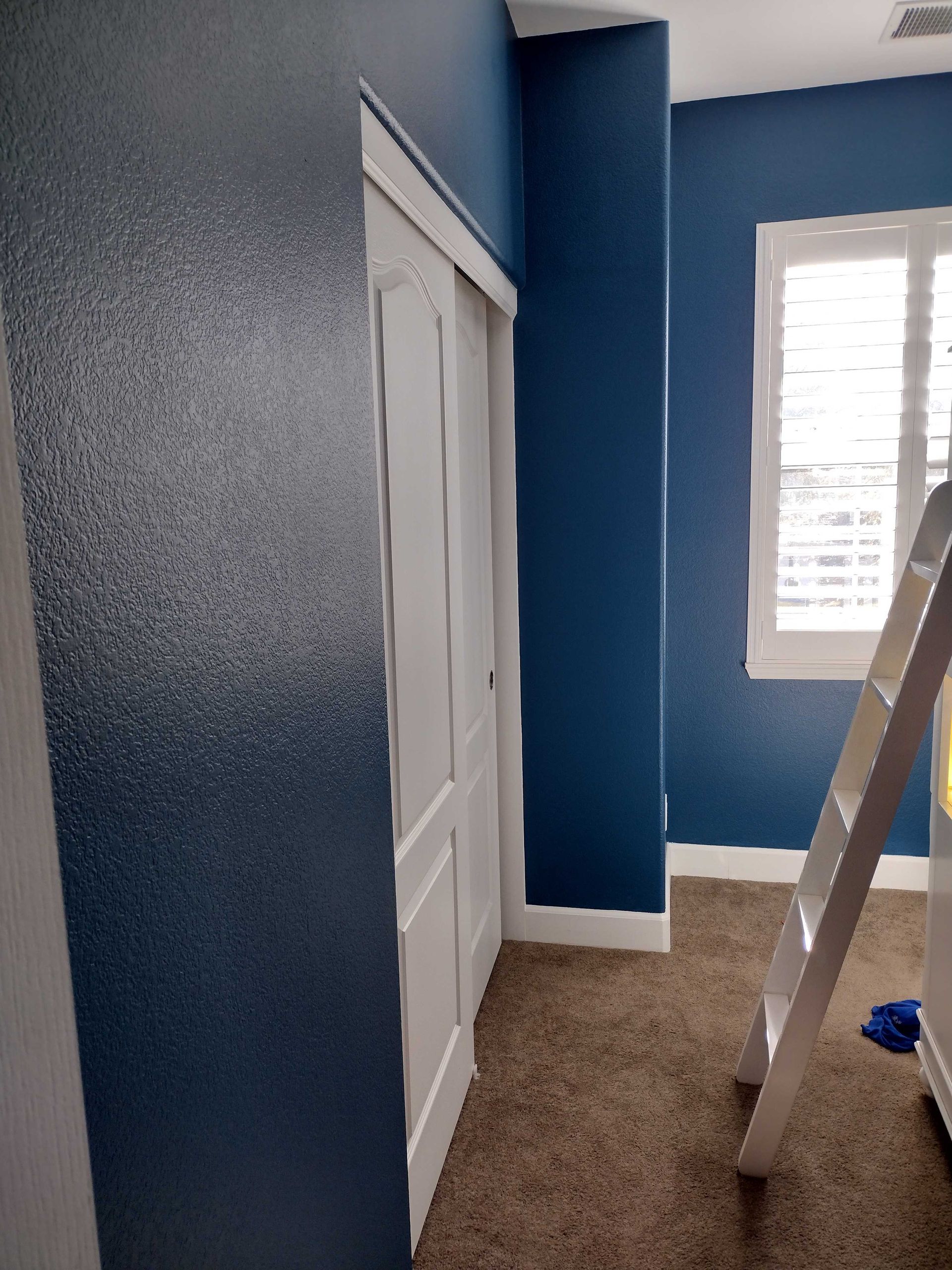 Blue bedroom under renovation with white closet doors, beige carpet, and a ladder by the window.