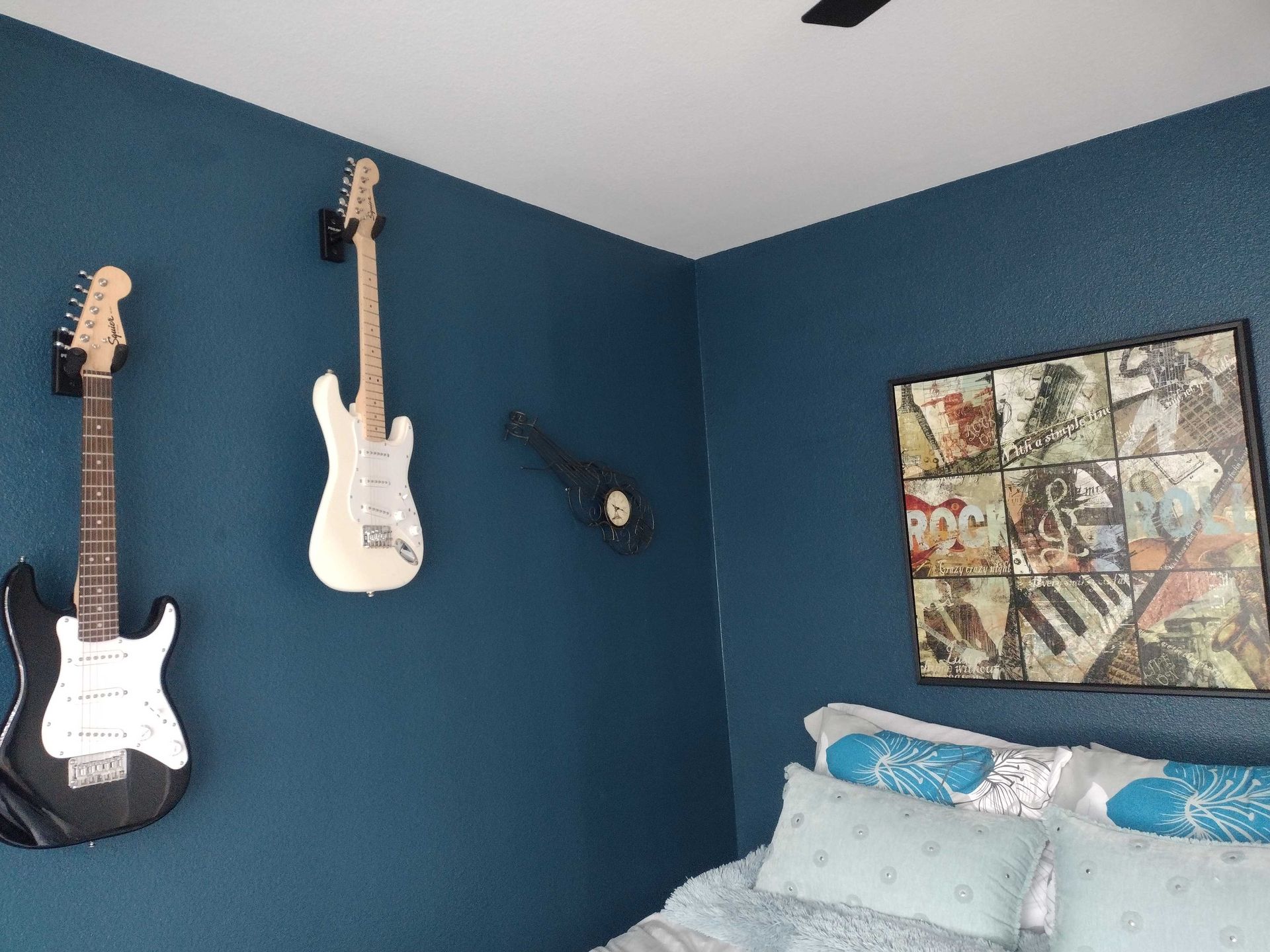 Blue bedroom corner with three guitars on the wall, a bed with blue pillows, and framed artwork.
