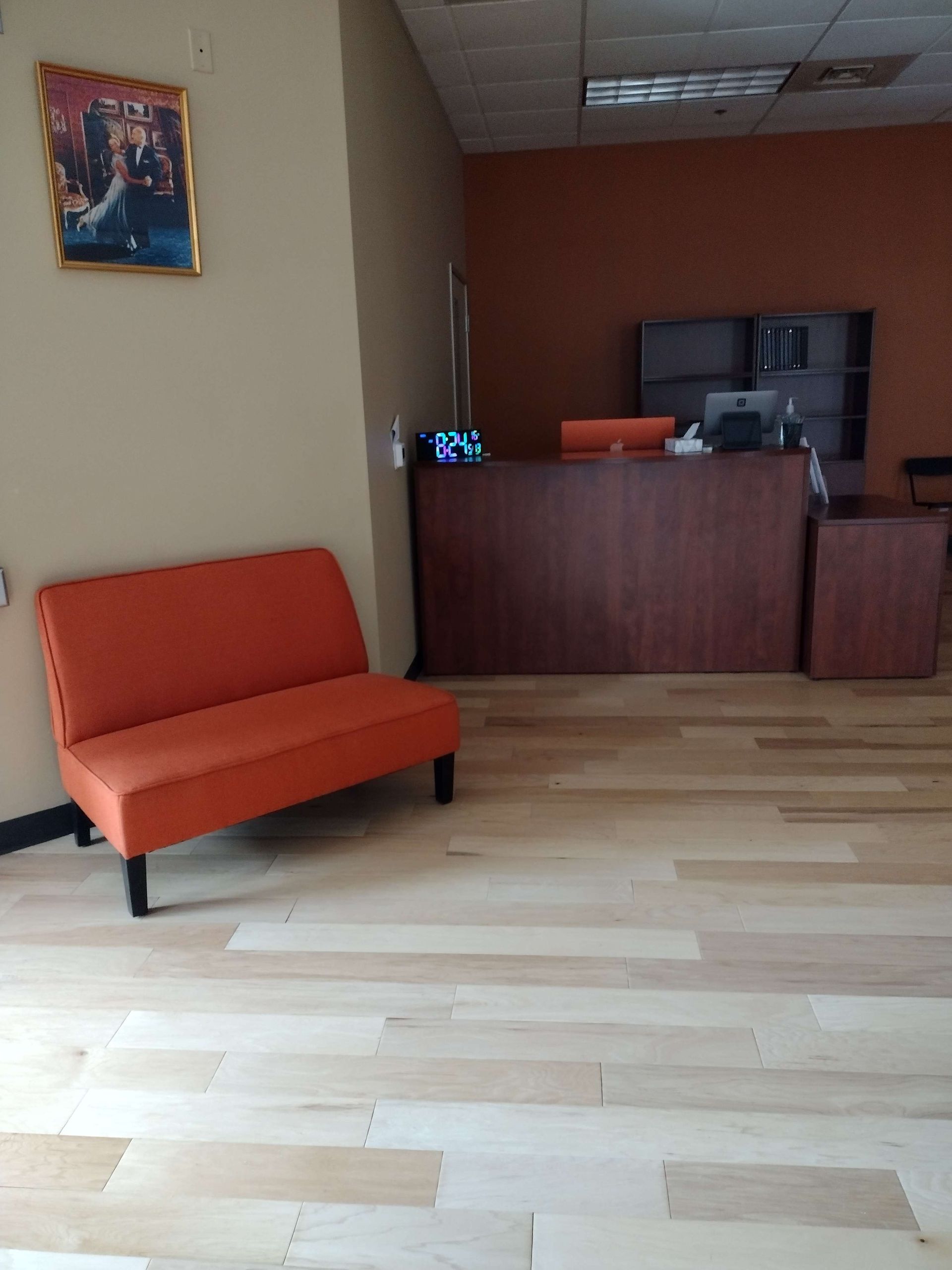 A waiting area with light wood flooring, an orange bench, a dark wood reception desk, and framed art on a beige wall.