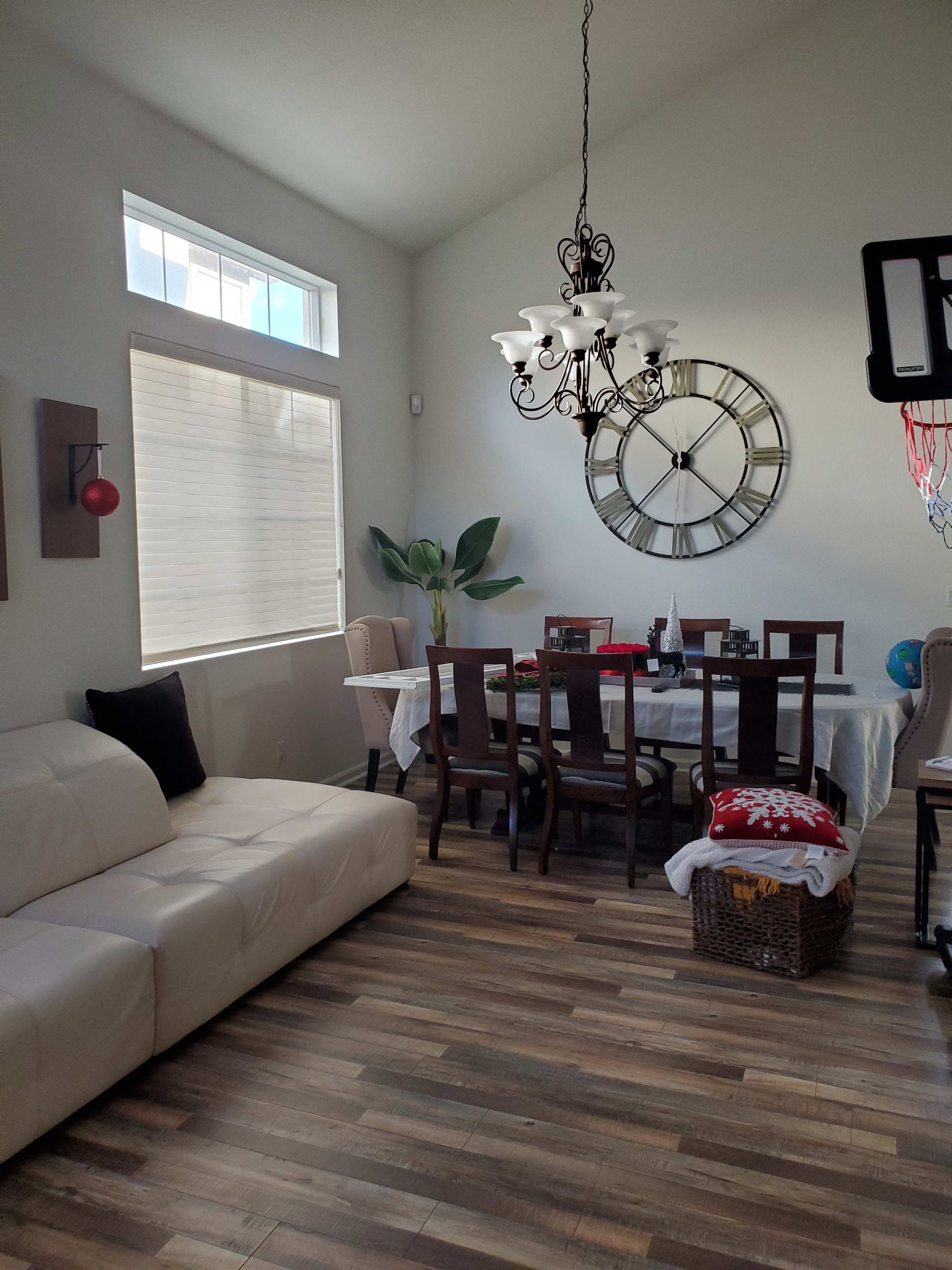 Cozy dining nook with beige sofa, wooden table and chairs, large wall clock, and tall windows with blinds.